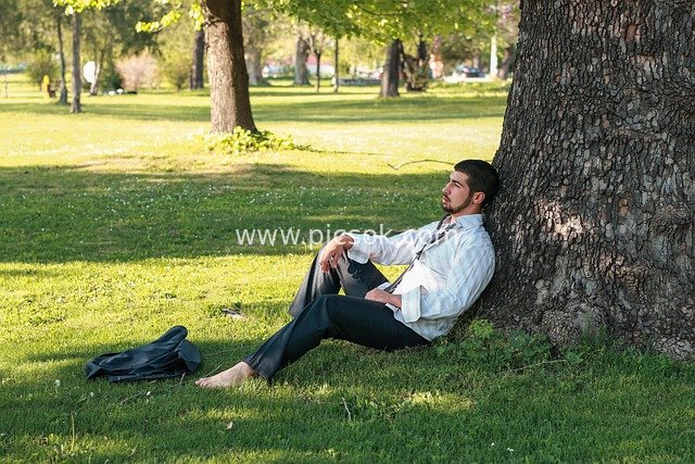 Young Man Relaxing on Park Lawn – Formal Attire, Barefoot, Leaning Against Tree