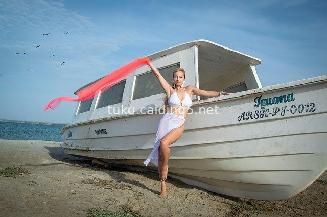 Bikini Model with Flowing Red Scarf by White Boat on Sandy Beach Under Blue Sky and Sea