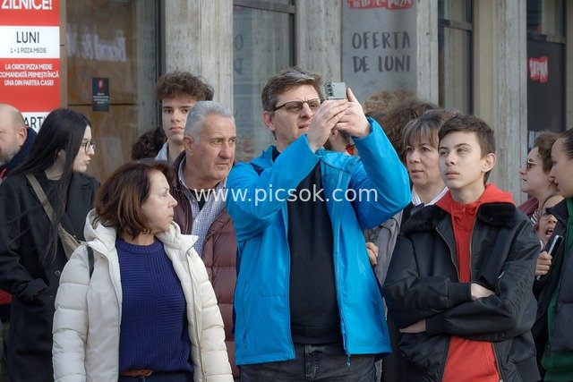 Crowd Gathers on City Street, Man Captures Moments with Smartphone