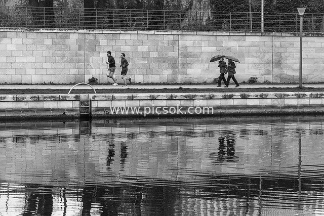 Black-and-White Image: Joggers and Umbrella-Carrying Pedestrians on a Waterfront Trail