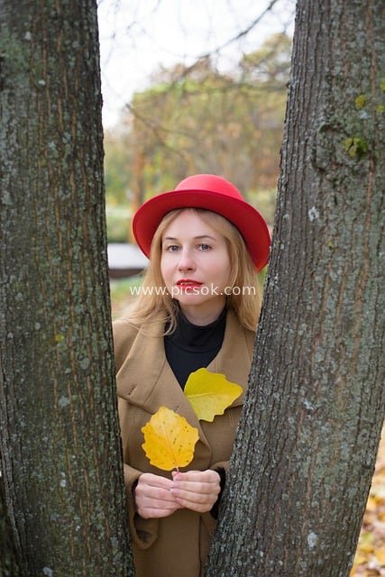 Autumn Fashion Portrait of a Gentle Young Woman in Red Hat and Camel Trench Coat in a Park with Fallen Leaves